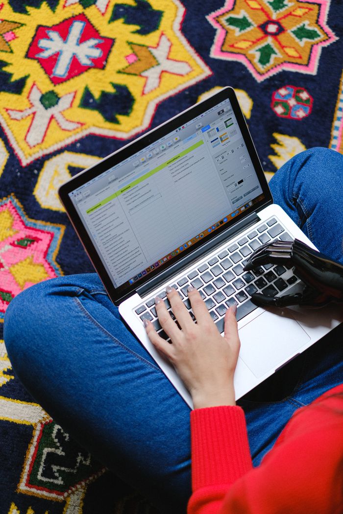 A person with a prosthetic hand typing on a laptop while sitting on a colorful patterned rug.